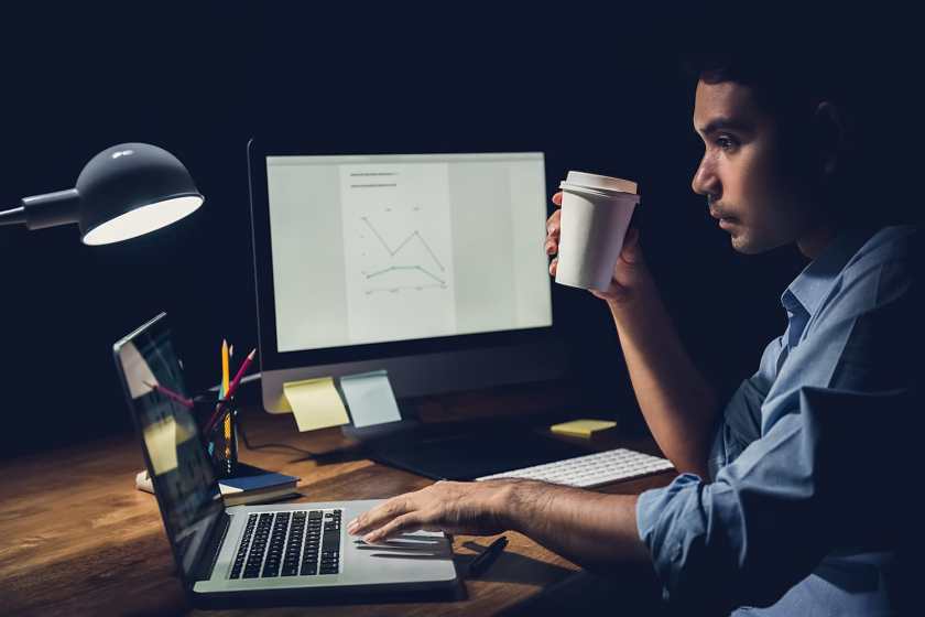 Sleepy tired businessman staying overtime late at night in the office holding coffee cup while focusing on working with laptop computer at his desk