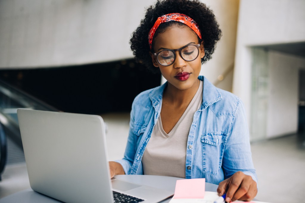 
Confident young African female entrepreneur sitting at a table in a modern office building lobby working on a laptop and reading notes in her planner