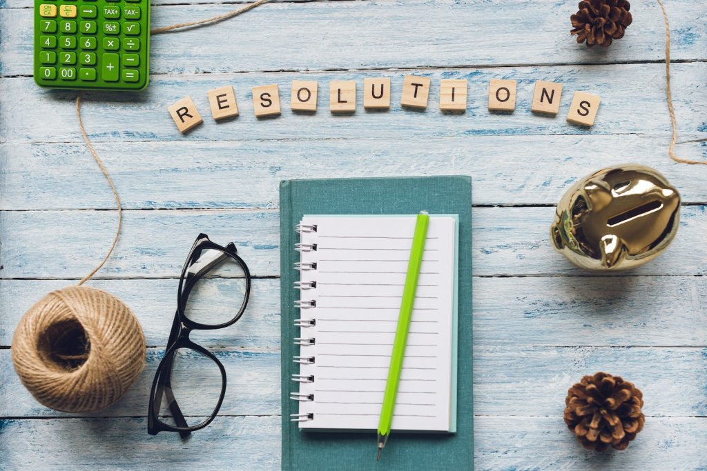 RESOLUTIONS word and an empty notepad, calculator, piggy bank, book and glasses on a blue and white wooden background. Top view, flat lay. New Year to do list concept.