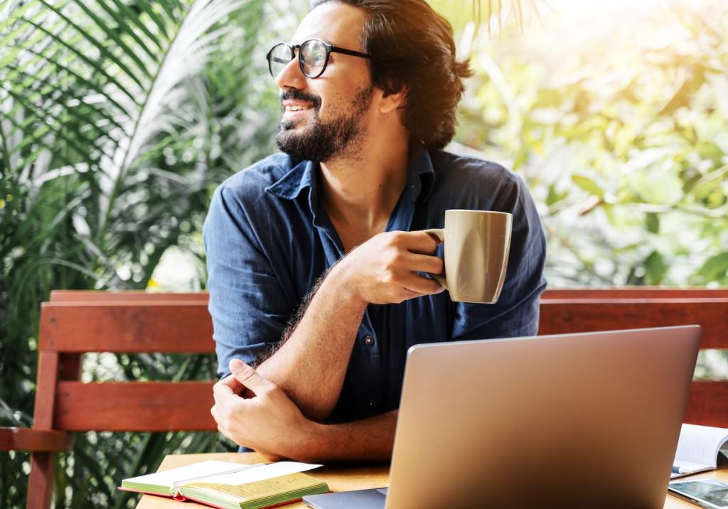 Handsome happy young curly latin man with a coffee mug in his hands sit on the terrace of the house in front of a laptop in the morning sunshine.