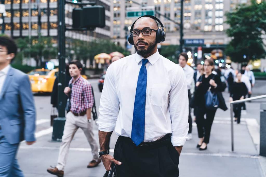 Formal bald and bearded black man in shirt and glasses using wireless headphones listening to music while walking against blurred crowded downtown looking away