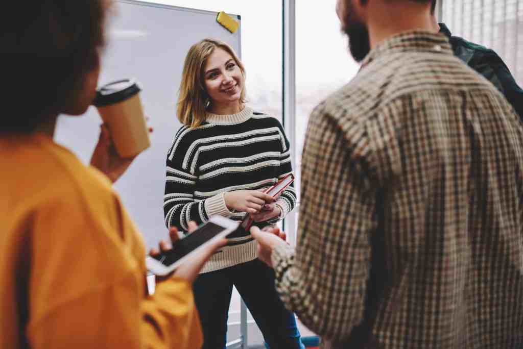 Positive young woman coach together with team of multicultural young people discussing productive strategy in front of whiteboard. Diverse workers dressed in casual wear communicating with each other