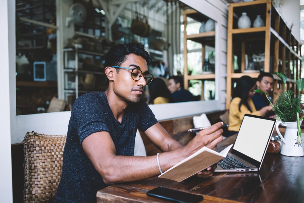 Thoughtful ethnic man in casual clothes and eyeglasses writing in planner while using laptop and sitting at table in tropical cafeteria