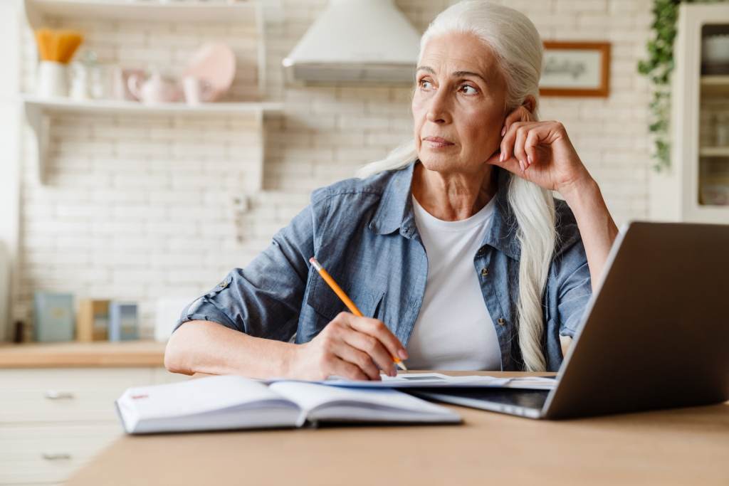 Pensive mature middle-aged woman working on computer while sitting at the table at home