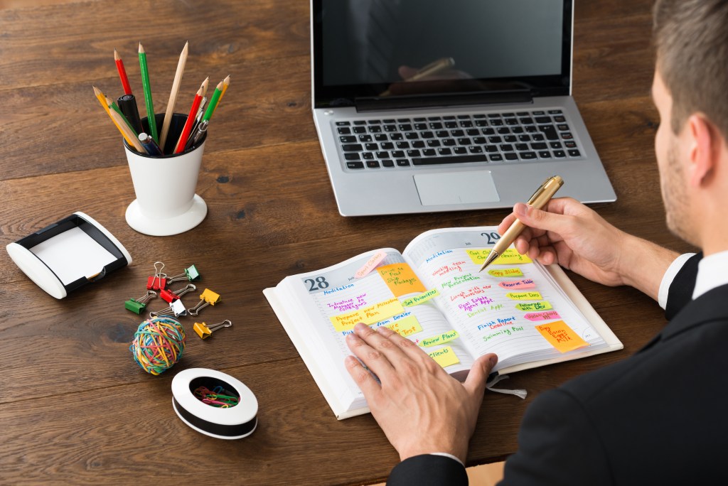 Close-up Of Businessman Writing Office Schedule In Diary With Stationery And Laptop At Desk