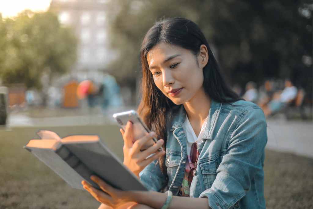 Young Asian woman reading a book but distracted by smarphone outdoors in the park