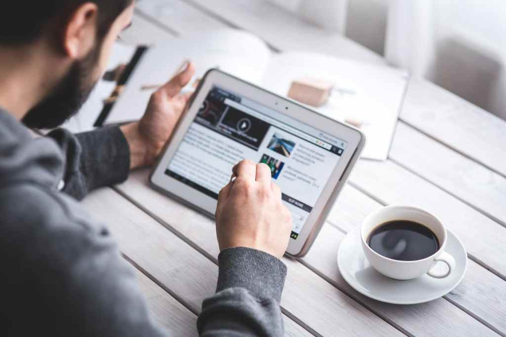 Man scrolling news website on ipad tablet device while drinking coffee